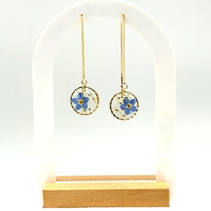 May include: A pair of gold dangle earrings with a small round pendant featuring pressed forget-me-not flowers.