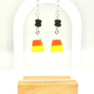 May include: A pair of Halloween-themed earrings. Each earring features a candy corn charm in yellow, orange, and white, topped with two black beads. The earrings hang from silver-colored hooks.