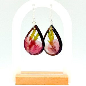 May include: A pair of teardrop-shaped earrings with a dark brown border. The earrings contain pink and yellow dried flowers encased in clear resin, suspended from silver-colored hooks. The earrings are displayed on a white stand with a wooden base.