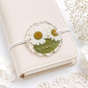 May include: A cream-coloured notebook secured with a white elastic band holding a circular, clear resin charm. The charm features two pressed white daisy flowers with yellow centres and green leaves, with the gold-coloured letters KMO embedded.