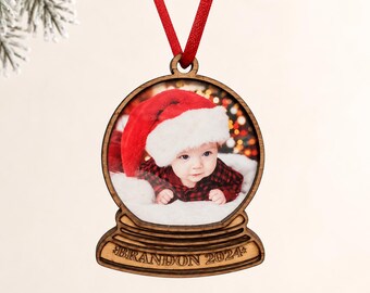May include: A wooden snow globe ornament featuring a photo of a baby wearing a red and white Santa hat and a red plaid shirt. The base of the ornament is engraved with the text BRANDON 2024 and it hangs from a bright red satin ribbon.
