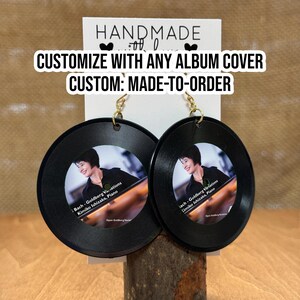 May include: A pair of black vinyl record earrings with gold hooks. The earrings feature a photo of a woman playing the piano with the text "Bach-Goldberg Variations Kimiko Ishizaka, Piano" printed on the front. The earrings are handmade and customizable with any album cover.