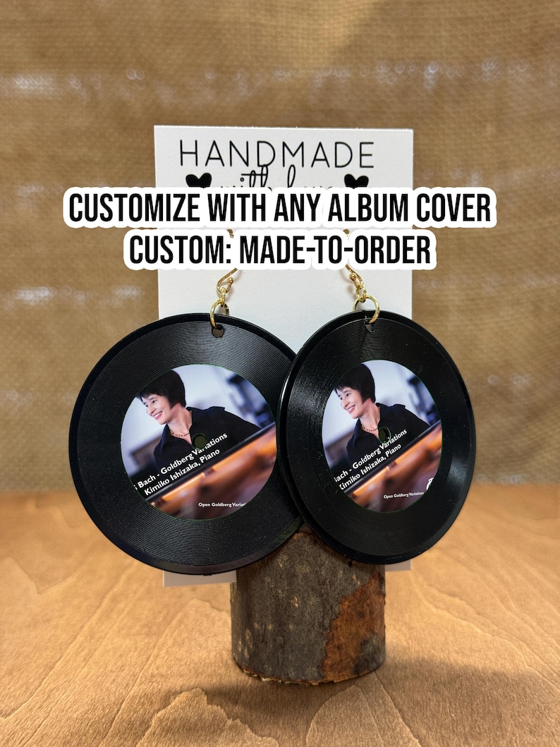 May include: A pair of black vinyl record earrings with gold hooks. The earrings feature a photo of a woman playing the piano with the text "Bach-Goldberg Variations Kimiko Ishizaka, Piano" printed on the front. The earrings are handmade and customizable with any album cover.