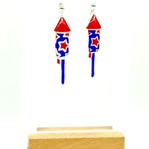 May include: A pair of patriotic earrings shaped like red, white, and blue fireworks. The earrings have a red top, a blue body with white stars, and a blue tail. They are displayed on a wooden stand against a white background.
