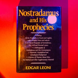 Puede incluir: Un libro negro con letras doradas titulado "Nostradamus y sus profecías". El libro incluye una biografía de Nostradamus, sus escritos y una bibliografía de sus comentaristas.
