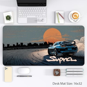 Pode incluir: Tapete de mesa com um carro desportivo azul à deriva numa estrada, com um pôr do sol e o horizonte da cidade ao fundo. A palavra "Supra" está escrita em branco. O tapete de mesa mede 40,6 x 81,3 cm.
