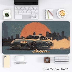 May include: A desk mat featuring a stylized illustration of a gray sports car with the word "Supra" in orange. The background includes a cityscape, a large orange sun, and a blue sky. The desk mat measures 16x32 inches.