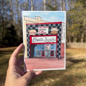 May include: Watercolor painting of Charlie Joseph's, a diner with red and black checkered accents. The sign reads "Charlie Joseph's Worlds Best Hot Dogs Hamburgers" and "Founded 1920". The image includes a parking sign.