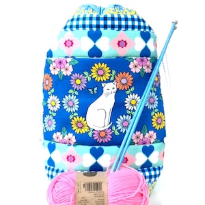 May include: A blue drawstring knitting project bag featuring a white cat illustration surrounded by colourful flowers. The bag includes a pink yarn ball and two blue knitting needles. The bag is decorated with floral and geometric patterns.