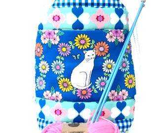 May include: A blue drawstring knitting project bag featuring a white cat illustration surrounded by colourful flowers. The bag includes a pink yarn ball and two blue knitting needles. The bag is decorated with floral and geometric patterns.