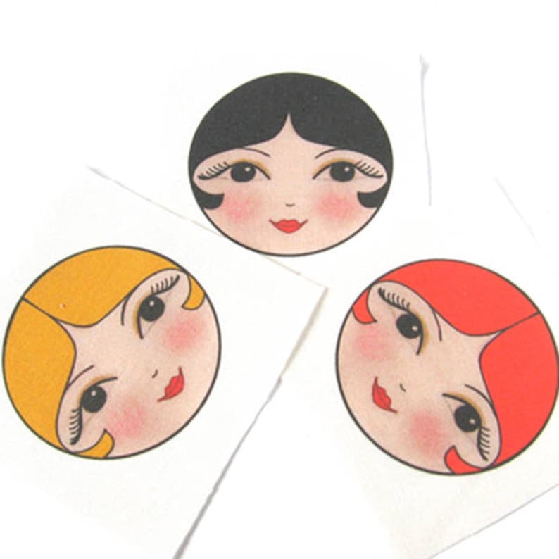 May include: Three round stickers with illustrations of women's faces. Each face has a different hair color: blonde, black, and red. The faces have black eyes, red lips, and rosy cheeks.