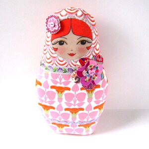May include: A pink and white fabric doll with a red hair and a pink flower on the chest. The doll is wearing a pink and orange patterned dress.
