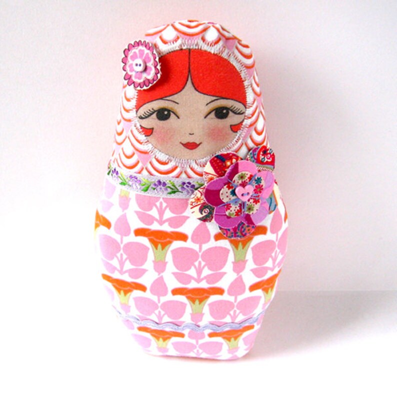 May include: A pink and white fabric doll with a red hair and a pink flower on the chest. The doll is wearing a pink and orange patterned dress.