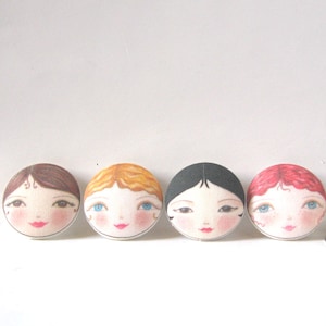 May include: Four round fabric buttons with painted faces of women. Each button features a different hairstyle and hair colour: brown, blonde, black, and red.