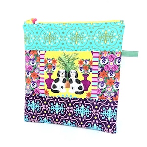 May include: A square, zippered pouch with a colourful patchwork design. The top section is turquoise with a repeating pattern. The middle section features a yellow background with two black and white dogs. The bottom section is purple with a teal pattern.
