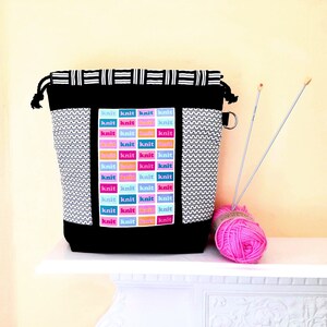 May include: Black drawstring bag with a white and black geometric pattern. The bag has a pocket with a colourful pattern of the word "knit" repeated. A pink ball of yarn and two knitting needles are in front of the bag.
