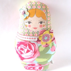 K&ouml;nnte beinhalten: Eine handgefertigte Stoffpuppe mit einem pink-, gr&uuml;n- und gelbfarbenen Blumenmuster. Die Puppe hat ein rundes Gesicht mit gelbem Haar, rosa Wangen und einem roten Mund. Die Puppe tr&auml;gt ein gr&uuml;n-wei&szlig; gepunktetes Kleid mit einer pinkfarbenen Blume an der Seite.