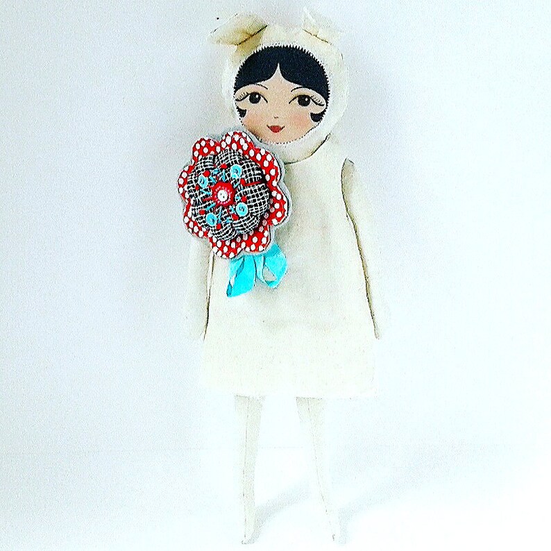 May include: A white doll wearing a white dress with a red and white flower pin. The doll has black hair and painted features.