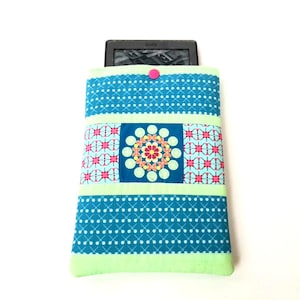 May include: A teal, light green, and pink quilted e-reader cover. The cover has a geometric pattern and a central square with a floral design. A pink button fastens the top flap. The cover is designed for an e-reader.