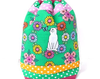 May include: A drawstring bag with a bright green background, decorated with a floral pattern and a white cat looking at a butterfly. The bag has pink, yellow, and white accents, creating a cheerful and whimsical design. The bag is made of fabric.