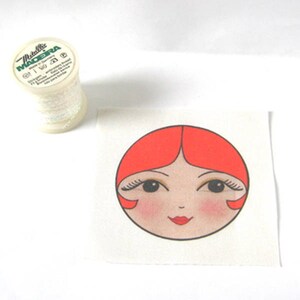 May include: A white fabric square with a red and white illustration of a woman's face. The woman has red hair, black eyelashes, and a red mouth.