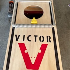 May include: A wooden game board with a black border and a red "V" logo. The board has a hole in the center and a spring-loaded trap door. The text "VICTOR" is printed on the board.
