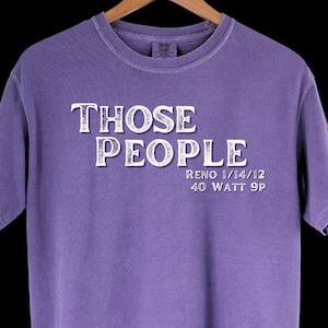 May include: A purple t-shirt with the words "THOSE PEOPLE" in white, block letters. Below the text are the words "RENO 1/14/12" and "40 WATT 9P" in white. The shirt is hanging on a wooden hanger.