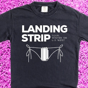 May include: A dark blue t-shirt with the text "Landing Strip" in white, followed by "Tour: Winter '06 U.S. West". A stylized image of a bikini bottom is below the text.