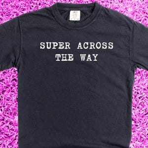 Pode incluir: Uma camiseta azul escura com texto branco que diz "SUPER ACROSS THE WAY".