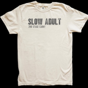 May include: Cream-colored t-shirt with the text "SLOW ADULT" in distressed gray lettering above "2ND STAGE CARNY". The shirt has a crew neck and short sleeves, and is made of a soft, comfortable material.