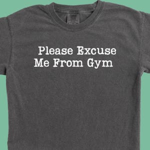 May include: Dark gray t-shirt with the text "Please Excuse Me From Gym" in white block letters. The shirt has a crew neck and short sleeves. The Comfort Colors brand tag is visible at the neckline.