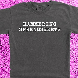 May include: A dark gray t-shirt with the text "HAMMERING SPREADSHEETS" printed in white.