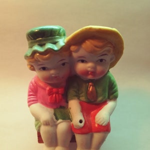May include: A pair of porcelain figurines of a boy and girl wearing hats and sitting side by side. The boy is wearing a yellow hat and a green shirt with red shorts. The girl is wearing a green hat and a pink shirt with green shorts.