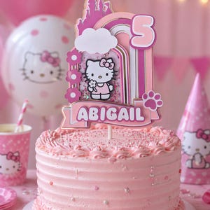 May include: A pink frosted cake with a Hello Kitty cake topper that reads "Abigail" and a number 5. The cake is decorated with sprinkles and sits on a white cake board. Party supplies, including balloons and cups, are in the background.