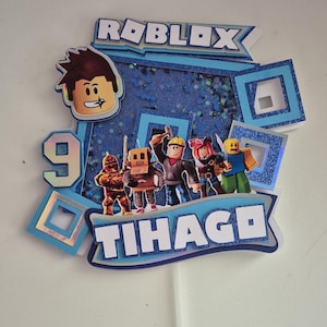 May include: A blue and white cake topper with the word "ROBLOX" and the name "TIHAGE" in a blue banner. The topper features a 9 and several Roblox characters, including a builder, a knight, a robot, a pirate, and a yellow character.