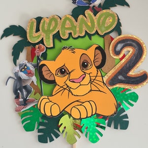 Könnte beinhalten: Ein Kuchentopper zum Thema König der Löwen mit Simba, Rafiki, Zazu und der Zahl 2. Der Topper ist aus Papier und hat ein grün-gelb-orangenes Farbschema.