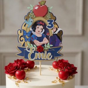 Op de afbeelding: Een wit geglazuurde taart versierd met rode rozen, rode appels en gouden accenten. Een taarttopper toont een Sneeuwwitje-ontwerp met de naam "Emie" en het nummer "3". De taart staat op een witte marmeren taartstandaard.