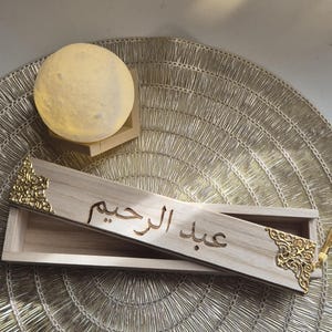 以下が含まれることがあります： アラビア書道と金色の装飾コーナーが付いた木製の箱。箱は開いており、内部が見えます。丸い、照らされた月の形をしたランプが、金色のテクスチャのある表面の背景にあります。