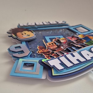 May include: A blue and white cake topper with a Roblox logo, a number 9, and the name "Tihana". The topper features a variety of Roblox characters, including a robot, a builder, and a knight.
