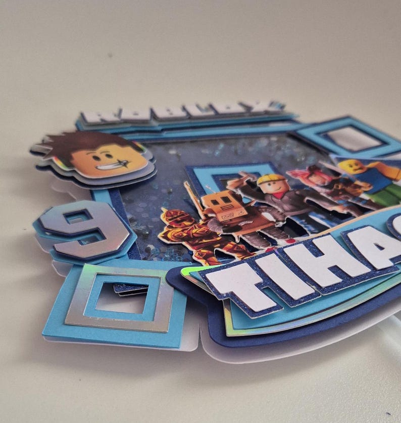 May include: A blue and white cake topper with a Roblox logo, a number 9, and the name "Tihana". The topper features a variety of Roblox characters, including a robot, a builder, and a knight.