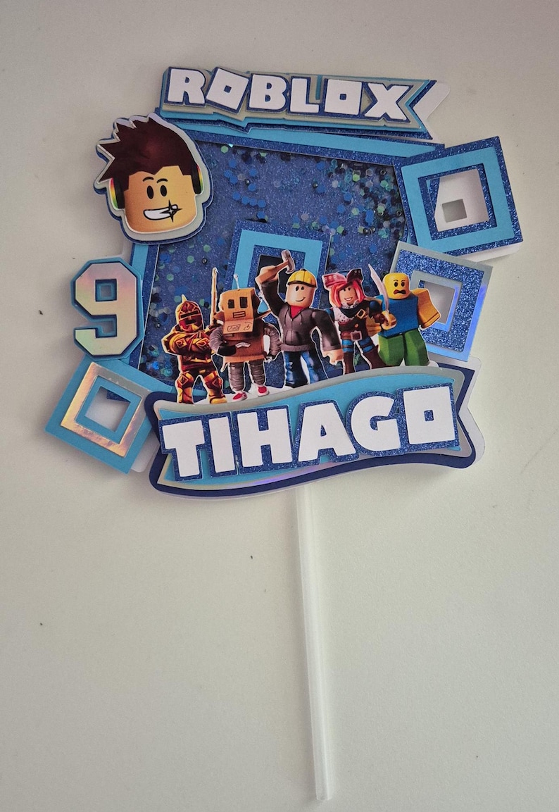 May include: A blue and white cake topper with the word "ROBLOX" and the name "TIHAGE" in a blue banner. The topper features a 9 and several Roblox characters, including a builder, a knight, a robot, a pirate, and a yellow character.