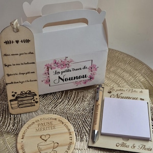 May include: A gift set comprising a white box with a handle, a wooden bookmark, a coaster, a notepad, and a pen. The box is decorated with floral accents and the text "Les petits trucs de Nounou". The bookmark and coaster have engraved text.