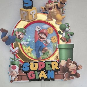 Peut inclure: Une décoration de gâteau multicouche représentant des personnages de Super Mario dont Mario, Luigi, Yoshi, Bowser, Donkey Kong et Toad. Le design comprend un cadre circulaire central avec un fond pailleté, des pièces d'or, une étoile et le texte SUPER GIAN.