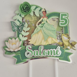 Könnte beinhalten: Kuchendekoration mit Prinzessin Tiana in einem grünen und gelben Kleid, einem Frosch und der Zahl 5. Der Aufsatz enthält einen Schmetterling, eine Blume und den Namen "Salomé" in weißer Schrift. Das Design ist mit grünem Glitzer akzentuiert.