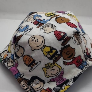 May include: A white fabric face mask with a colorful Peanuts cartoon print featuring characters like Charlie Brown, Snoopy, Lucy, and Linus.