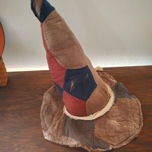 May include: A tall, pointed hat constructed from quilted fabric patches in shades of brown, red, and dark blue. The hat features a wide, circular brim and a frayed, off-white fabric trim around the base of the crown, with visible stitching.