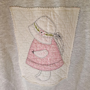 May include: A light gray heathered sweatshirt featuring a quilted cream-colored patch on the front. The patch displays an embroidered figure wearing a bonnet and a pink floral dress with a white bird held in the hands, outlined in black stitching.
