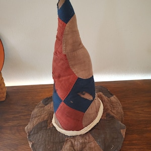 May include: A tall, pointed hat constructed from a patchwork of fabric pieces in shades of red, navy blue, and brown. The conical crown features visible frayed edges and small tears, while the wide, circular brim is finished with a white trim.