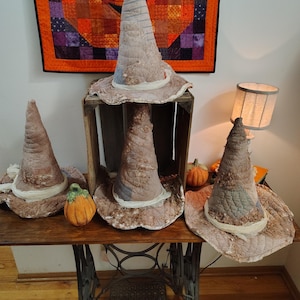 May include: Four fabric witch hats in shades of brown, tan, and grey with textured, quilted surfaces and frayed edges. The hats feature pointed, conical crowns and wide, floppy brims. Two small, decorative orange pumpkins are placed near the hats.
