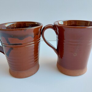 May include: Two handmade ceramic mugs in shades of brown. The mugs have a rustic, earthy appearance with a smooth, glossy finish. One mug has dark brown drips. The mugs have a handle and a slightly flared rim.